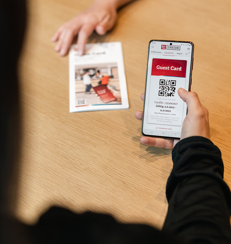 Eine Person hält ein Smartphone in der Hand, auf dem die digitale Gästekarte Ötztal mit einem QR-Code abgebildet ist, während eine andere Person auf der anderen Seite des Tisches ein Papierdokument hält. Beide sitzen an einem hellen Holztisch.