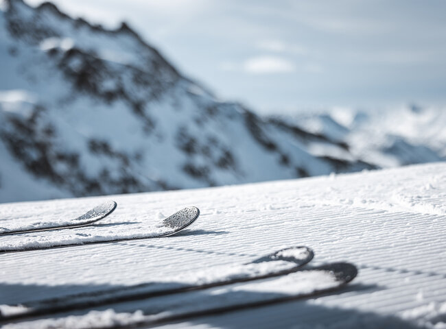 Die Skier ruhen auf dem präparierten Schnee, während die verschneiten Berggipfel im Hintergrund verschwimmen.