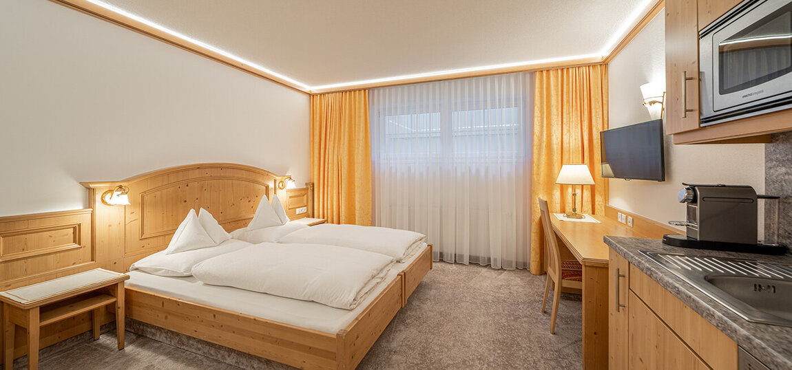 Ein gemütliches Apartmentzimmer mit einem Doppelbett, Holzmöbeln, einem Schreibtisch mit einer Lampe, einem an der Wand befestigten Fernseher und einer Küchenzeile. Die warme Beleuchtung und die gelben Vorhänge vor dem Fenster schaffen eine einladende Atmosphäre.