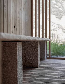 Eine Nahaufnahme einer minimalistischen Holzbank mit Steinauflagen, die auf einer Holzterrasse neben vertikalen Holzlatten steht und einen Blick auf Grünflächen und eine verschwommene Landschaft im Freien bietet.