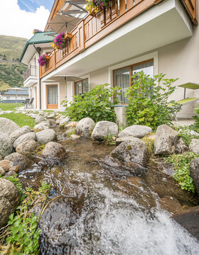 Ein kleiner Bach fließt über Felsen neben einem Gebäude, dessen Balkone mit bunten Blumen geschmückt sind; im Hintergrund sind Berge und ein teilweise bewölkter Himmel zu sehen.