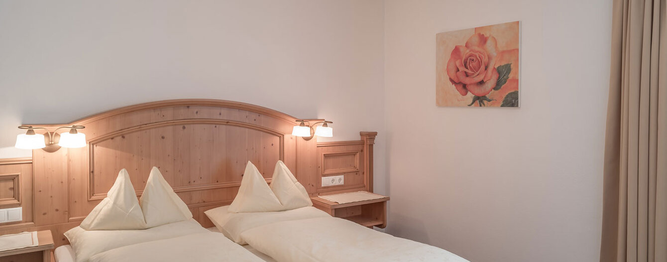 Ein gemütliches Hotelzimmer mit einem hölzernen Kopfteil, Nachttischen mit Lampen, beiger Bettwäsche, einem Rosenbild an der Wand und beigen Vorhängen.