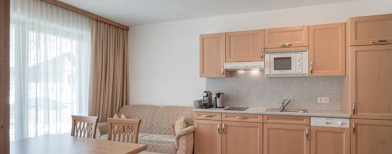 Ein helles, modernes Apartment mit hellen Holzschränken, einer kleinen Küchenzeile, einem beigen Sofa, einem Esstisch mit Stühlen, einem großen Fenster mit Vorhängen und neutral getönten Wänden und Fußböden.