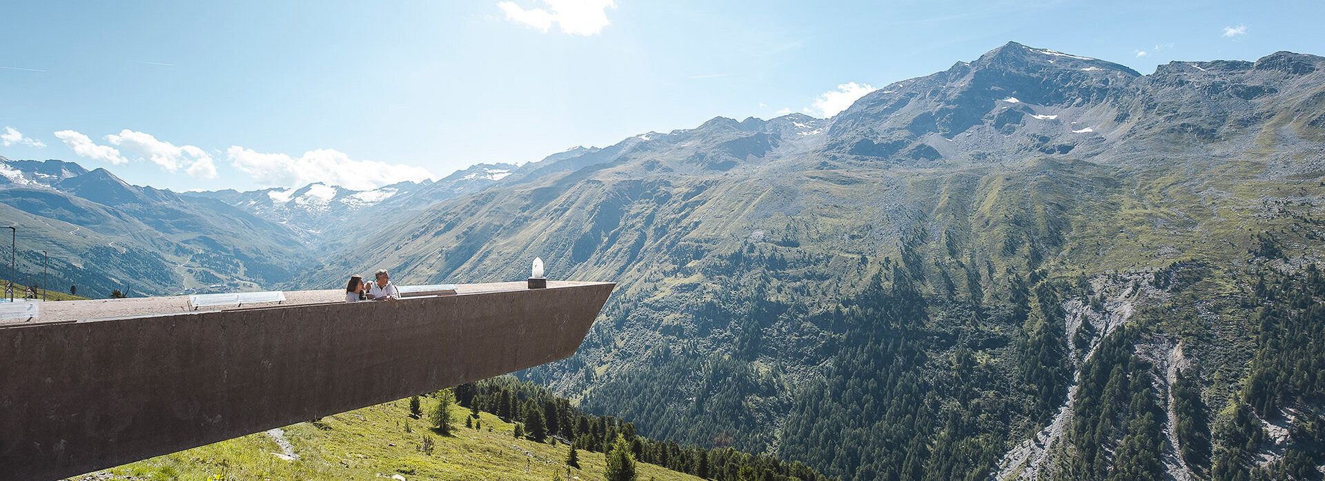 Eine moderne Aussichtsplattform ragt über ein üppiges, grünes Bergtal hinaus, auf der Menschen stehen, umgeben von hohen Bergen unter einem klaren blauen Himmel.