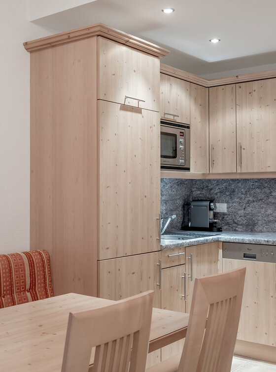 Eine moderne Küche mit hellen Holzschränken, grauen Arbeitsplatten, Einbaugeräten und Deckenbeleuchtung. Im Vordergrund steht ein Esstisch aus Holz mit passenden Stühlen und einer gepolsterten Bank.