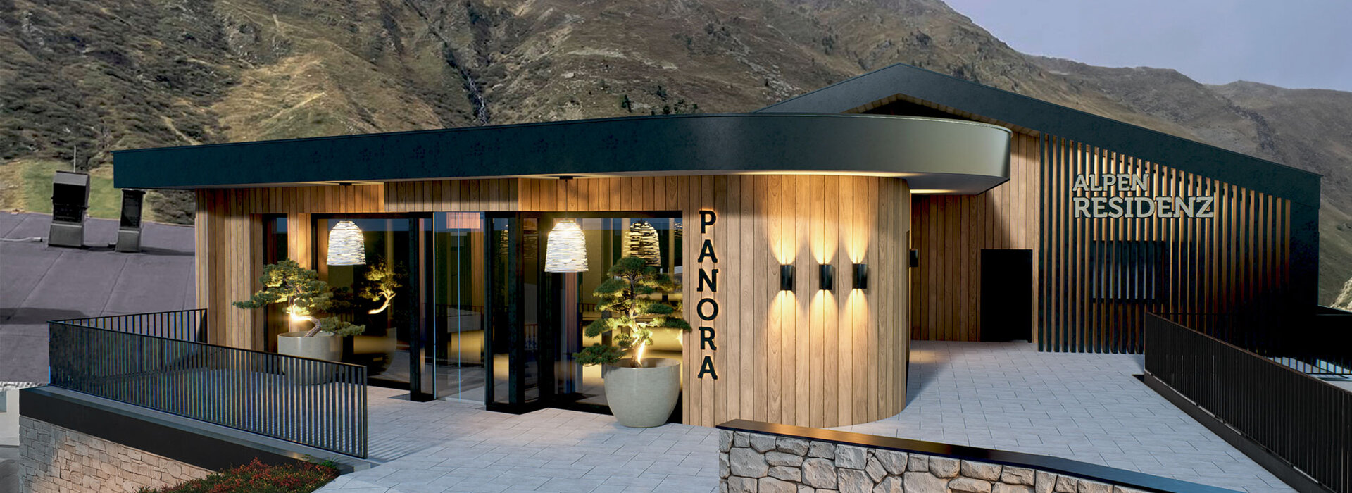 Ein modernes Gebäude aus Holz und dunklem Metall mit der Aufschrift PANORA an der Vorderseite und ALPENRESIDENZ an der Seite, vor dem Hintergrund felsiger, grasbewachsener Berge unter einem wolkenverhangenen Himmel.