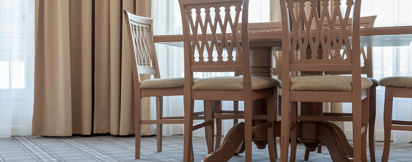 Vier Holzstühle stehen um einen Esstisch in einem Raum mit raumhohen beigen Vorhängen und großen Fenstern. Der Boden ist mit einem grau-karierten Teppich ausgelegt.