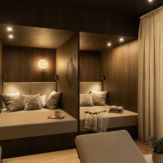 Zwei gemütliche, holzgetäfelte Sitzecken mit gedämpfter Beleuchtung, beigen Kissen, einer Decke, gestapelten Büchern und einer Tasse, getrennt durch eine Trennwand, mit einem hellen Vorhang auf der rechten Seite.