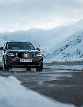 Ein schwarzer BMW-Geländewagen fährt auf einer klaren Bergstraße, umgeben von schneebedeckten Gipfeln und unter einem bewölkten Himmel.
