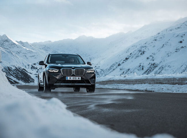 Ein schwarzer BMW-Geländewagen fährt auf einer klaren Bergstraße, umgeben von schneebedeckten Gipfeln und unter einem bewölkten Himmel.