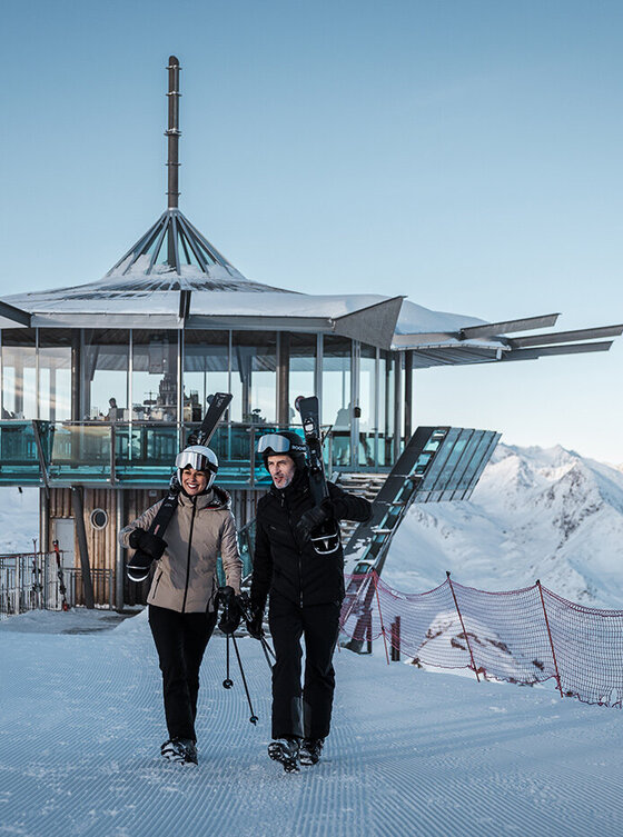 Zwei Skifahrer in Winterausrüstung stehen vor einer modernen gläsernen 360 Grad Bar mit verschneiten Bergen im Hintergrund.