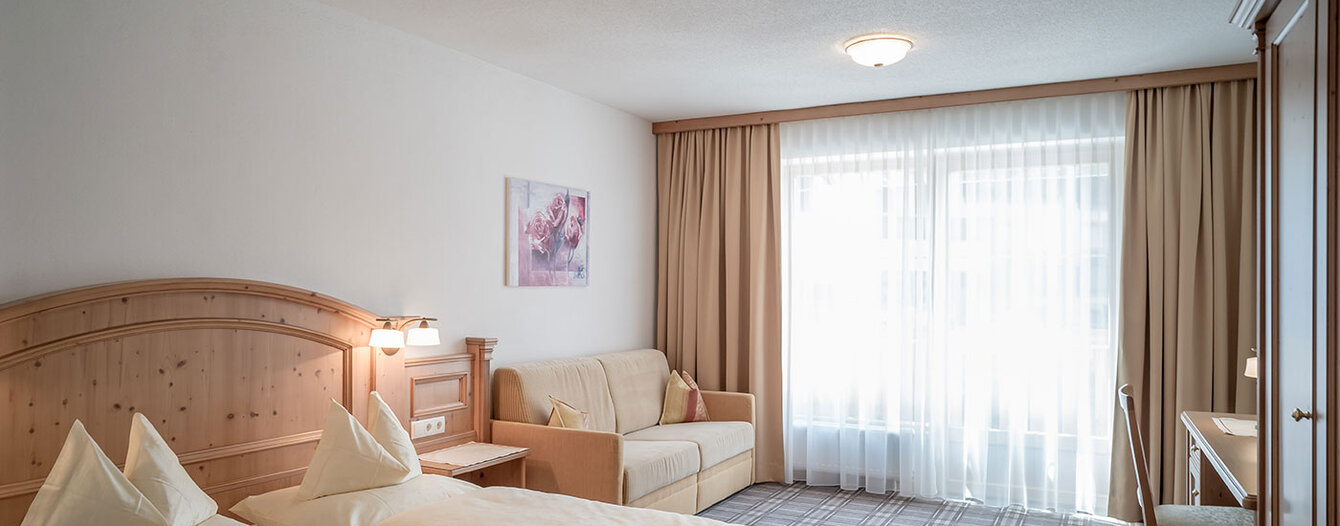 Ein helles Apartmentzimmer mit einem Doppelbett, einem hölzernen Kopfteil, einem gelben Sofa, einem Schreibtisch mit Stuhl, einem großen Kleiderschrank und einem Fenster mit durchsichtigen Vorhängen, die das Tageslicht hereinlassen.