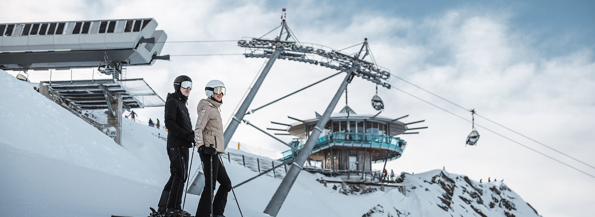 Zwei Personen in Skiausrüstung stehen auf einer verschneiten Piste in der Nähe eines Skilifts und von Seilbahnen, mit einem modernen Berggebäude und blauem Himmel im Hintergrund.