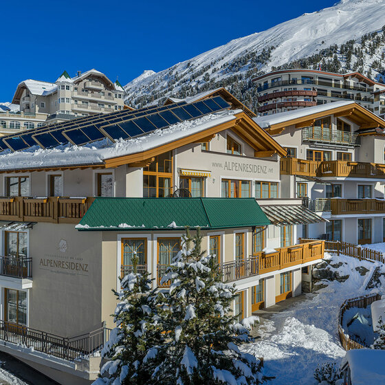 Ein modernes Alpenhotel mit Photovoltaikanlagen auf dem Dach ist von schneebedeckten Gebäuden und Bäumen umgeben, vor der Kulisse verschneiter Berghänge und eines strahlend blauen Himmels. 