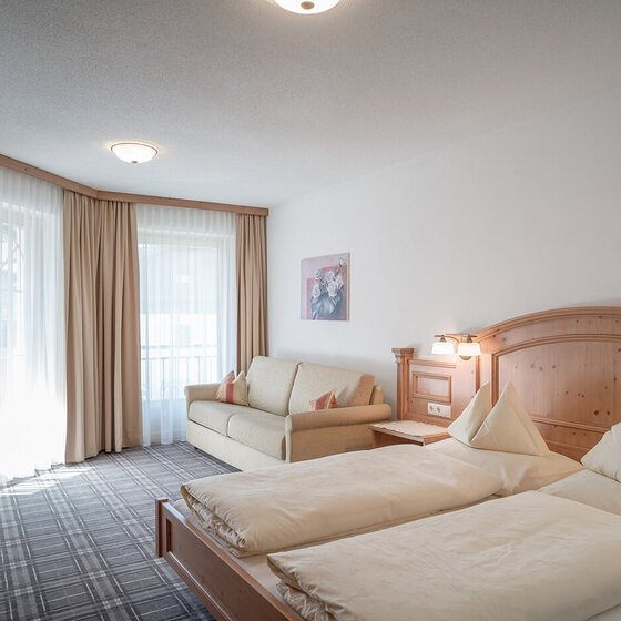 Ein helles Apartmentzimmer mit ordentlich gemachtem Bett, einem beigen Sofa, Holzmöbeln, einem karierten Teppich, hellen Vorhängen und sanftem Tageslicht, das durch große Fenster fällt.