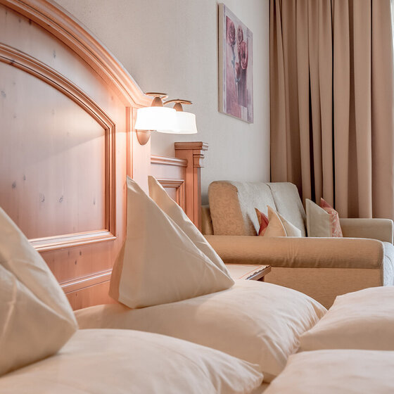 Ein gemütliches Apartmentzimmer verfügt über sauber gemachtes Bett mit weißer Bettwäsche, ein hölzernes Kopfteil, ein beiges Sofa mit dekorativen Kissen, eine Wandlampe, Vorhänge und ein Fenster, das natürliches Licht hereinlässt.