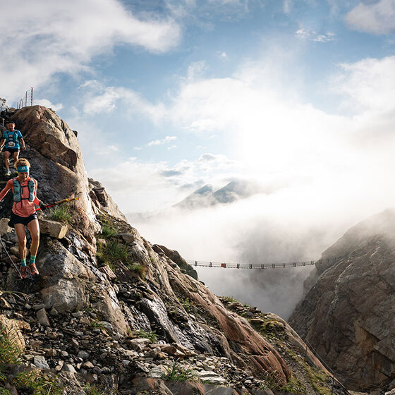 Zwei Trailrunner in Sportkleidung steigen einen felsigen Bergpfad mit saftig grünem Gras hinab. Nebel und Wolken füllen die Täler, und eine Hängebrücke überspannt die Klippen im Hintergrund unter einem strahlenden Himmel.
