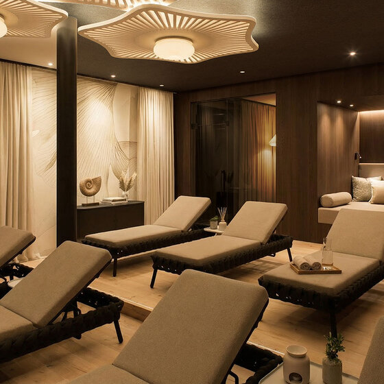 Ein moderner Ruheraum mit gepolsterten Liegen, sanfter Beleuchtung, hölzernen Akzenten und gemütlicher Einrichtung. Handtücher und Getränke stehen auf Tabletts bereit, und im Hintergrund befinden sich private Sitzbereiche, die eine entspannte Atmosphäre schaffen.