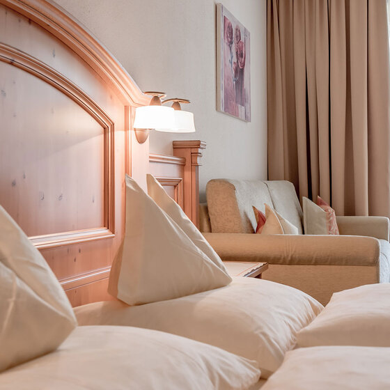 Ein helles Hotelzimmer mit ordentlich gemachtem Bett, einem hölzernen Kopfteil, einem cremefarbenen Sofa mit Kissen, beigen Vorhängen, einer Wandlampe und einem gerahmten Bild über dem Sofa neben einem Fenster mit durchsichtigen Vorhängen.