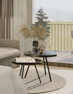Eine gemütliche Wohnzimmerszene mit zwei runden schwarzen Beistelltischen auf einem runden Teppich, dekoriert mit einer Vase mit getrockneten Blumen, Glaswaren und Dekorationsgegenständen, neben einem Sofa und einem großen Fenster mit Blick auf einen Balkon und die Berge.