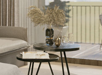 Eine gemütliche Wohnzimmerszene mit zwei runden schwarzen Beistelltischen auf einem runden Teppich, dekoriert mit einer Vase mit getrockneten Blumen, Glaswaren und Dekorationsgegenständen, neben einem Sofa und einem großen Fenster mit Blick auf einen Balkon und die Berge.