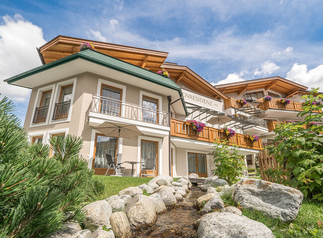 Ein modernes Alpenhaus mit Holzbalkonen und bunten Blumen, umgeben von Grün und Felsen mit einem kleinen Bach im Vordergrund, unter einem strahlend blauen Himmel mit vereinzelten Wolken.