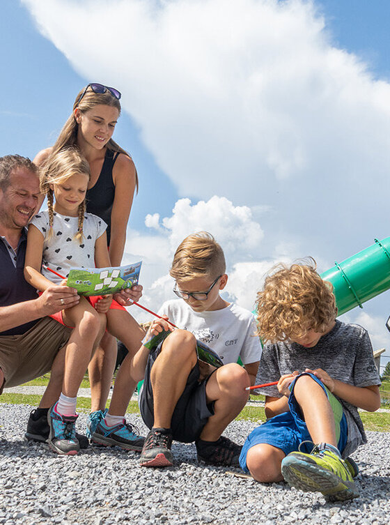 Eine fünfköpfige Familie schaut sich im Freien gemeinsam eine Karte an, wobei zwei Erwachsene und drei Kinder lächeln. Hinter ihnen steht der Spielplatz vom Widiversum, eine große Comic-Schafsfigur mit einer grünen Rutsche vor einem hellen, teilweise bewölkten Himmel.