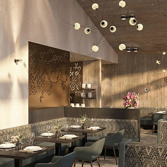 Ein modernes Restaurant mit gemusterten Stühlen, Holzwänden und -decken, schwarzen Tischen, grünen Stühlen, großen Fenstern, Hängelampen und dekorativer Wandkunst. Auf einem Tresen im hinteren Bereich sind rosa Blumen arrangiert.
