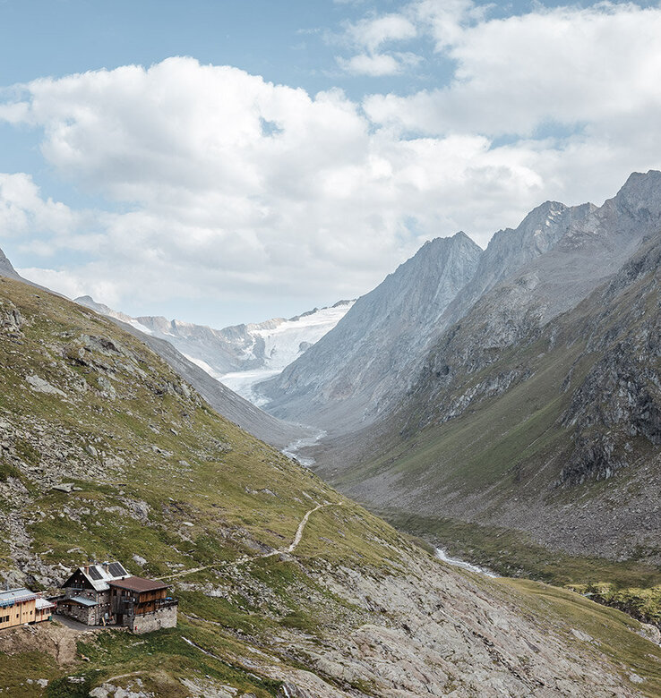 Gebirgstal mit einem kleinen Holzhaus, felsigen Hängen und einem Gletscher in der Ferne unter einem teilweise bewölkten Himmel.