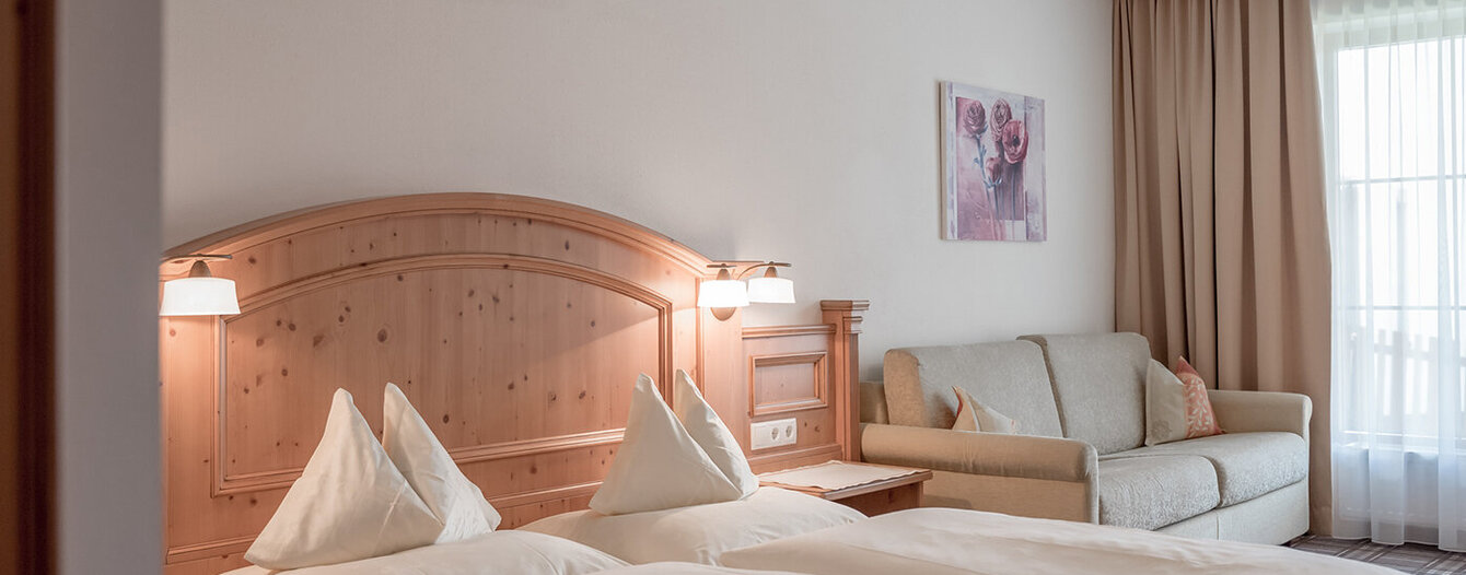 Ein gemütliches Apartmentzimmer mit sauber gemachtem Bett, einem hölzernen Kopfteil, sanfter Beleuchtung, einem beigen Sofa, einem Fenster mit Vorhängen und einem Blumengemälde an der Wand.