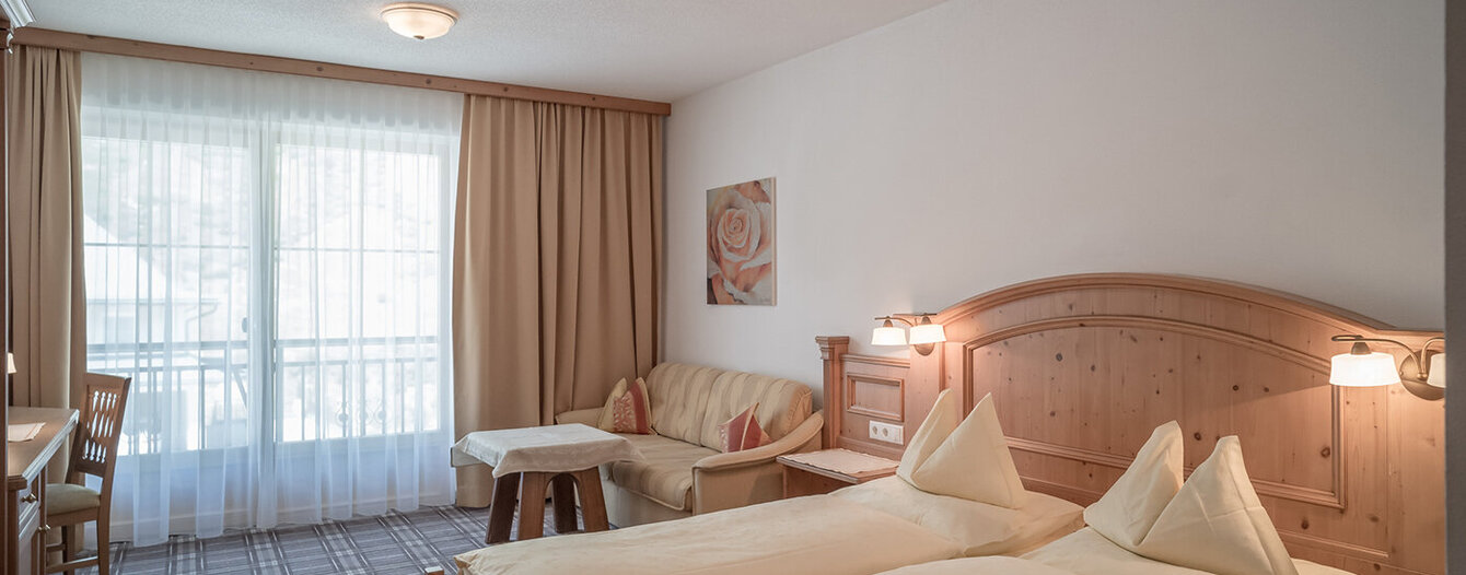 Ein gemütliches Apartmentzimmer mit sauber gemachtem Bett, einem hölzernen Kopfteil, einem Sofa, einem kleinen Tisch, einem Schreibtisch mit einem Stuhl und großen Fenstern mit durchsichtigen Vorhängen, die das Tageslicht hereinlassen.