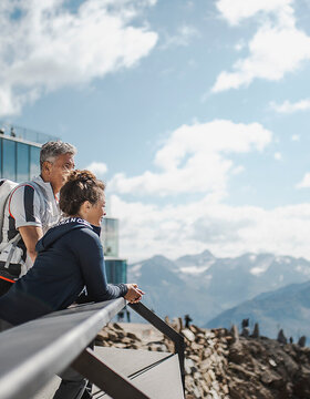 Zwei Personen stehen an einem Geländer mit Blick auf die Berge und ein modernes Gebäude unter einem teilweise bewölkten Himmel.