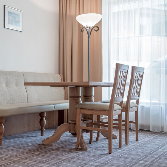 Ein heller Raum mit einem Holztisch, zwei passenden Holzstühlen, einer gepolsterten Bank, einer Stehlampe und großen Fenstern, die mit durchsichtigen Vorhängen und beigen Gardinen bedeckt sind.