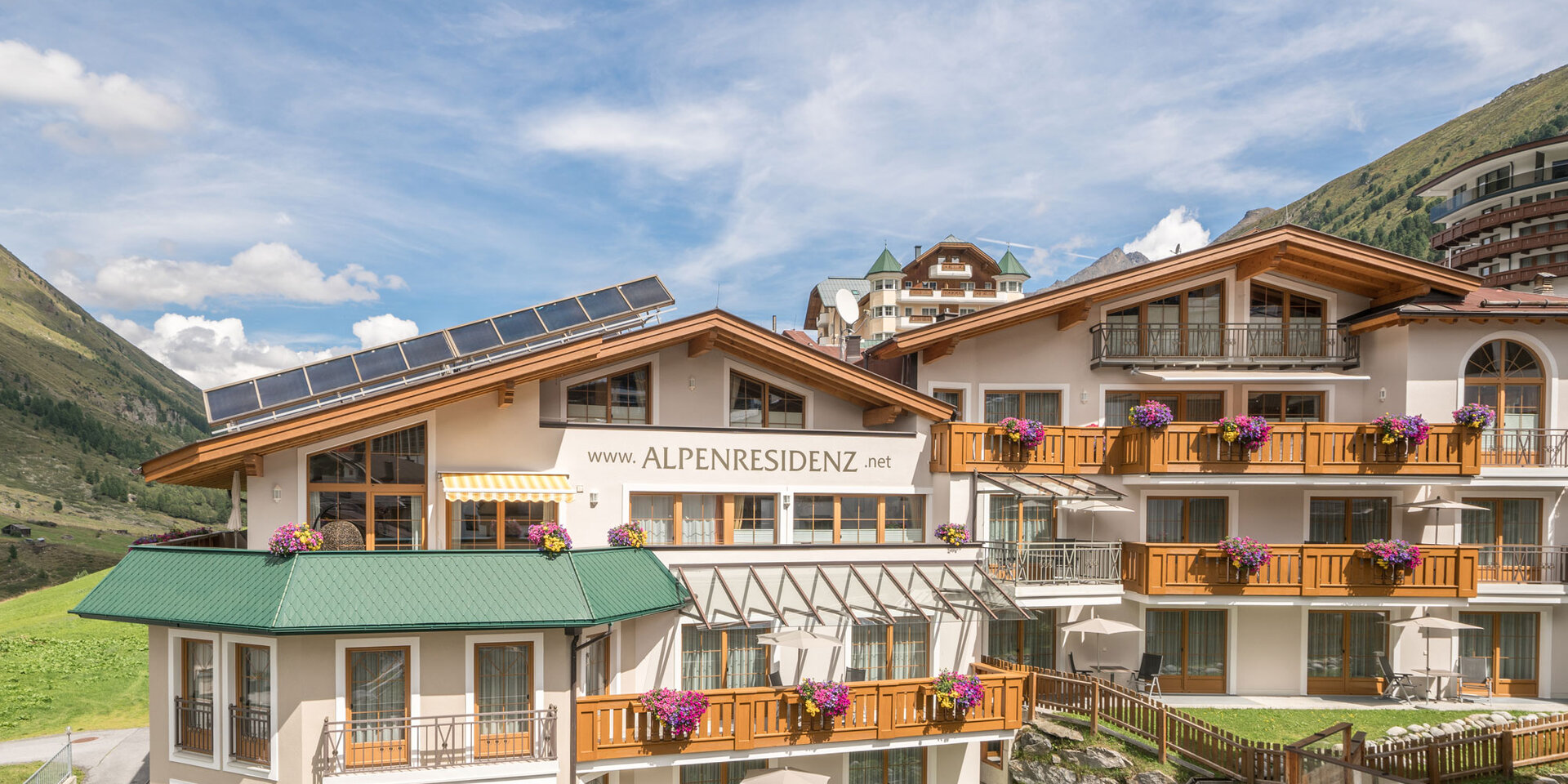 Gezeigt wird die Alpenresidenz mit Balkonen und Blumenkästen in einer grünen Berglandschaft unter einem blauen Himmel mit vereinzelten Wolken.