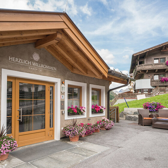 Gemütlicher Gebäudeeingang mit Holztür, Blumenkästen und einem Schild mit der Aufschrift Herzlich Willkommen. Sitzbereich im Freien mit Stühlen und einem Tisch auf der rechten Seite. Im Hintergrund ist ein weiteres blumengeschmücktes Gebäude zu sehen.