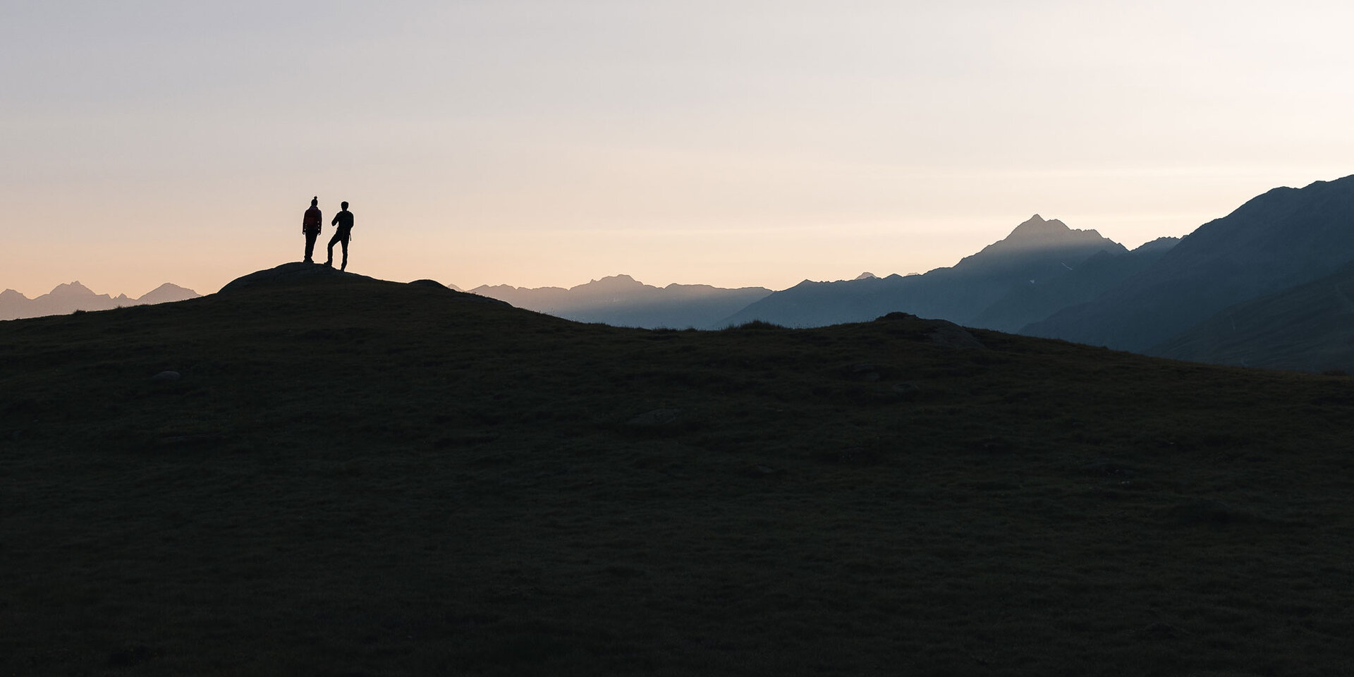 Zwei Personen stehen auf einer Bergkuppe vor einem pastellfarbenen Sonnenuntergangshimmel mit Bergen im Hintergrund. Die Szene vermittelt ein Gefühl von Frieden, Abenteuer und natürlicher Schönheit.