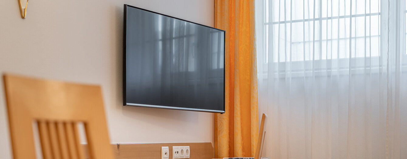 Ein modernes Apartmentzimmer verfügt über einen an der Wand montierten Flachbildfernseher über einem Holztisch mit Stuhl. Ein Fenster mit weißen Gardinen und orangefarbenen Vorhängen lässt natürliches Licht herein.