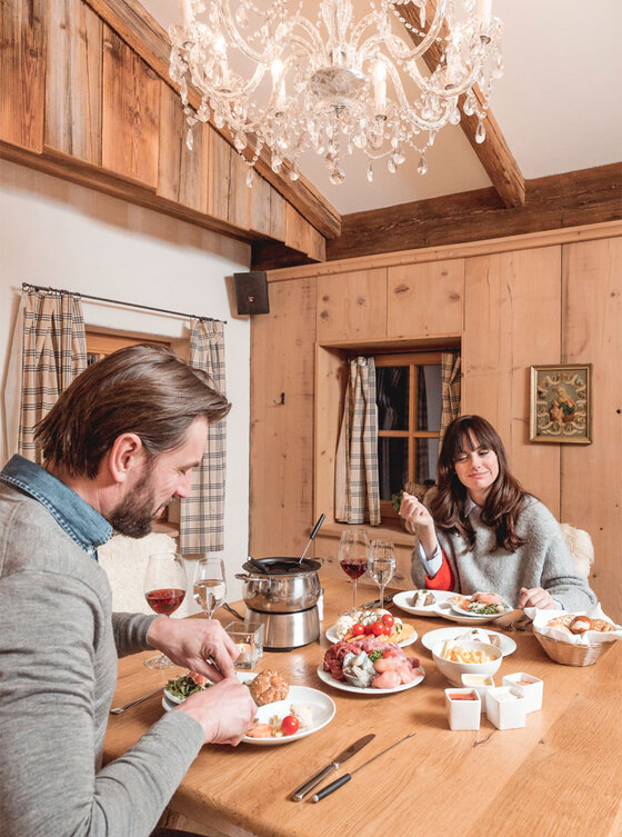 Ein Mann und eine Frau genießen gemeinsam eine Fondue-Mahlzeit an einem hölzernen Esstisch in einem gemütlichen, rustikalen Raum mit einem Kronleuchter.