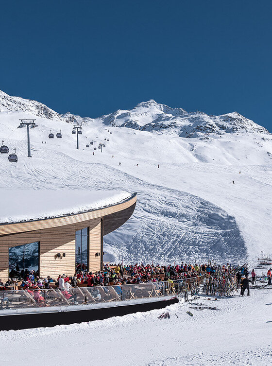 Skifahrer versammeln sich vor einer modernen Holzhütte auf einem verschneiten Berg mit Skiliften und strahlend blauem Himmel.