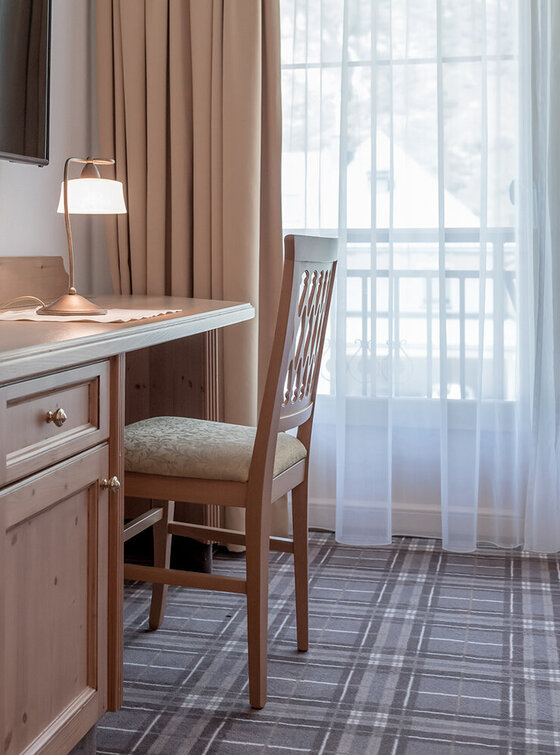 Ein kleiner Holztisch mit einem Stuhl, einer Schreibtischlampe und einem montierten Fernseher steht in einem gemütlichen Zimmer mit Teppichboden an einem Fenster mit durchsichtigen Vorhängen. Natürliches Licht fällt durch die beigen Vorhänge.