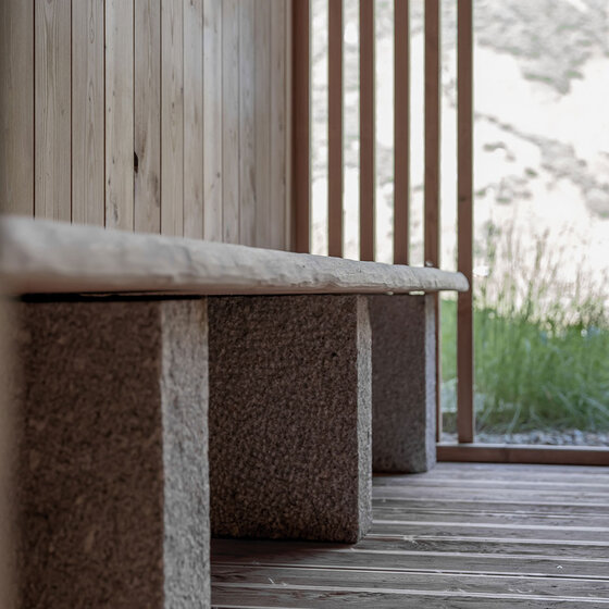 Eine Holzbank mit steinernen Stützen steht auf einem Holzboden neben vertikalen Holzlatten, während das Sonnenlicht die natürliche Umgebung im Hintergrund beleuchtet.