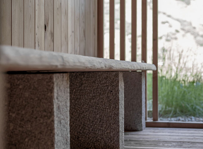 Eine Holzbank mit steinernen Stützen steht auf einem Holzboden neben vertikalen Holzlatten, während das Sonnenlicht die natürliche Umgebung im Hintergrund beleuchtet.