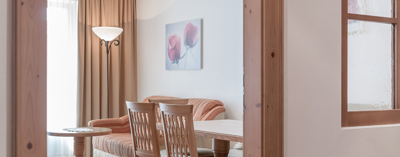 Ein gemütliches Wohnzimmer mit einem beigen Sofa, einem Holztisch und Stühlen, einer Stehlampe und einem Gemälde mit roten Blumen an einer weißen Wand. Durch ein Fenster mit beigen Vorhängen fällt Licht ein.