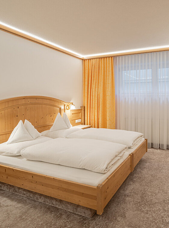 Ein gemütliches Apartmentzimmer mit einem Doppelbett, Holzmöbeln, einem Schreibtisch mit Lampe, einem an der Wand montierten Fernseher und einer kleinen Küchenzeile. Große Fenster mit gelben Vorhängen lassen natürliches Licht herein und schaffen eine warme Atmosphäre.